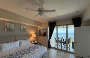 Ein Hotelzimmer mit Bett, Deckenventilator, Nachttisch mit Lampe, Bildern an den Wänden und einem Balkon mit Meerblick.