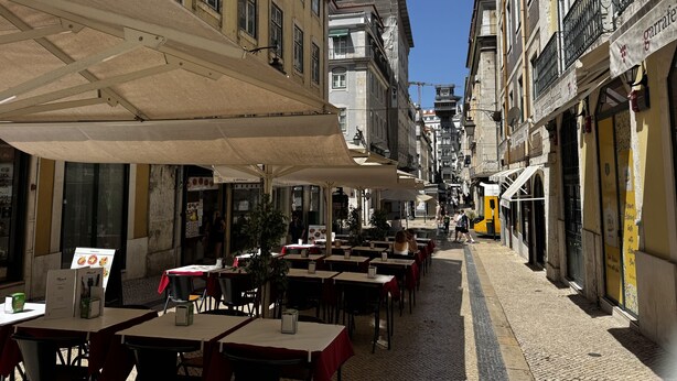 A narrow street lined with outdoor dining tables and umbrellas, with buildings on either side. - Lux Old Town (Lisboa)