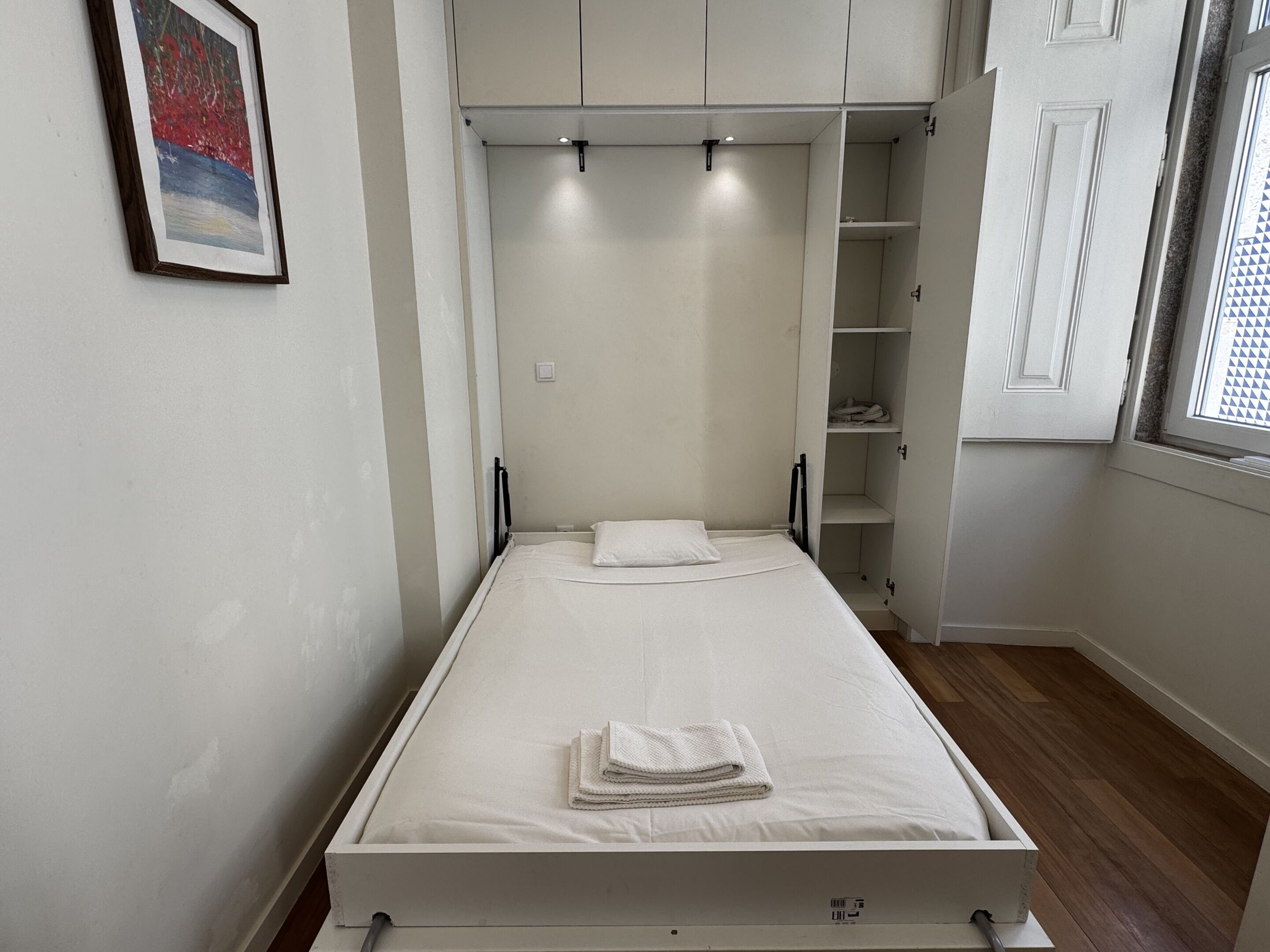 A single bed with a white mattress and pillows, a small bedside table, a wardrobe with open doors, and a window with a geometric pattern.