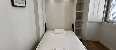 A single bed with a white mattress and pillows, a small bedside table, a wardrobe with open doors, and a window with a geometric pattern.