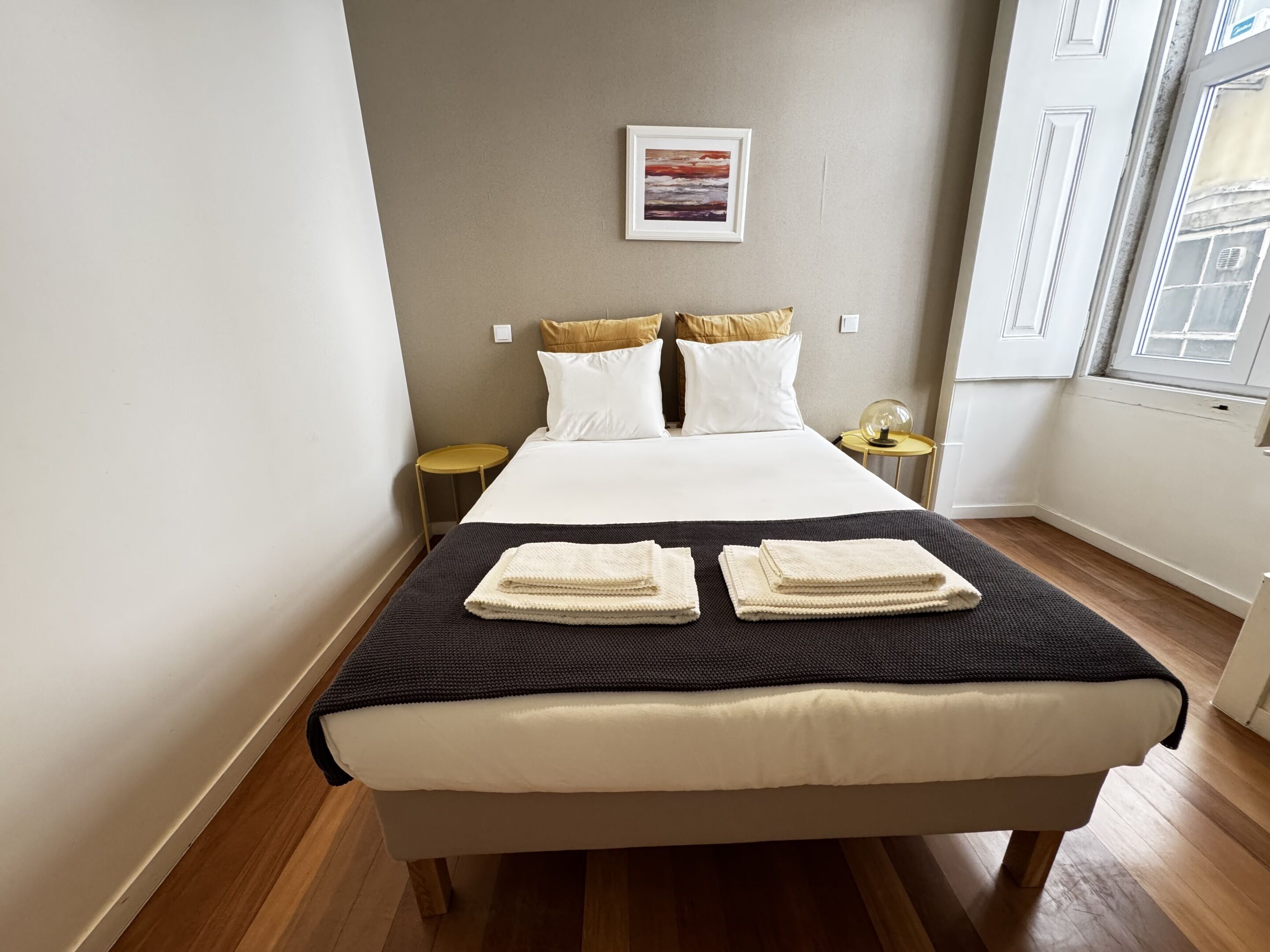 A neatly made bed with white linens and two folded towels, a small round table with a lamp, and a framed picture on the wall.