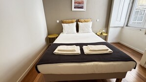 A neatly made bed with white linens and two folded towels, a small round table with a lamp, and a framed picture on the wall. - Lux Old Town (Lisboa)