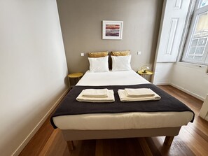 A neatly made bed with white linens and two folded towels, a small round table with a lamp, and a framed picture on the wall. - Lux Old Town (Lisboa)