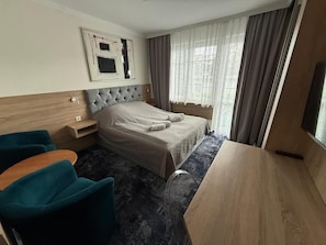 Ein Hotelzimmer mit einem Bett, einem Schreibtisch, zwei Stühlen, einem Nachttisch und einem Vorhangfenster.