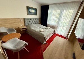 Ein Hotelzimmer mit einem Bett, zwei Stühlen, einem kleinen Tisch, einem Holzkopfende, einem Bild an der Wand und einem Fenster mit Vorhängen.