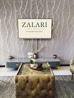 Uma sala de estar moderna com uma parede cinza estampada, um letreiro emoldurado com a inscrição 'ZALARI LUXE SUITES BY ZALARI LUXE STAYS', um sofá azul com pés amarelos, um banquinho estofado e uma mesa de centro com objetos decorativos.