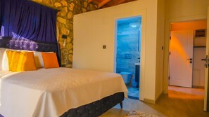 Ein Schlafzimmer mit einem Bett, einer Steinwand, einem durch eine offene Tür einsehbaren Badezimmer und einer Tür, die in einen weiteren Raum führt.