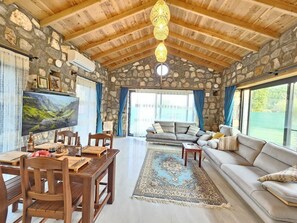 Ein rustikales Interieur mit Steinwänden, Holzbalken, einem Essbereich mit Tisch und Stühlen, einer Couch und einem großen Fernseher.
