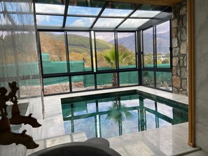 A modern interior with a glass-enclosed pool, stone walls, and a view of a mountainous landscape. - Luxurious 2-bedroom stone villa with two pools, Turkish hamam & sauna (İncirköy)