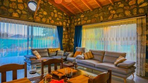 Una sala de estar acogedora con paredes de piedra, dos sofás, una mesa de comedor con sillas y ventanas grandes con cortinas.