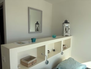 Una cama con estructura blanca y cabecera de consola, un espejo encima, dos repisas con objetos decorativos y una mesita de noche con lámpara.