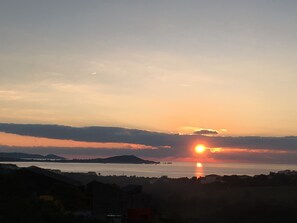 Un coucher de soleil sur une étendue d’eau, avec des reliefs et des bâtiments visibles au loin.