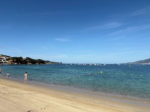 A beach with clear blue water, a sandy shore, and a distant view of boats and buildings.