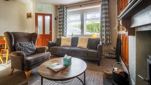 Een woonkamer met een bank, fauteuil en salontafel, een open haard en een raam met gordijnen.
