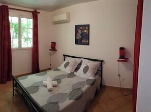Une chambre à coucher avec un lit, une fenêtre avec des rideaux, un climatiseur, une œuvre d’art accrochée au mur et une table de chevet avec une lampe.