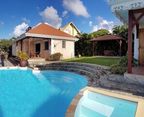 Une piscine bordée de pierres, une maison avec un toit rouge et un patio couvert.
