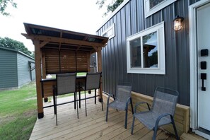 A covered outdoor seating area with a table and chairs, a wooden deck, and a building with a window and exterior wall. - Less House, More Lake | Perfect for Couples (Kingston)