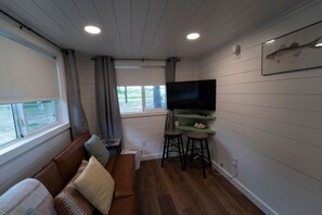 A compact living space with a sofa, a TV mounted on a wall, and a window with curtains. - Less House, More Lake | Perfect for Couples (Kingston)