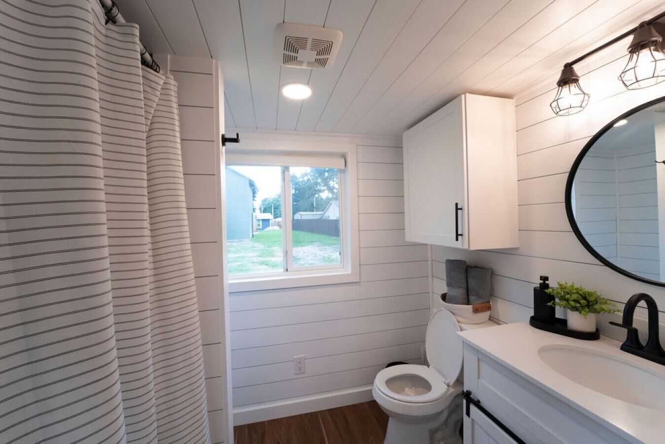 Ein Badezimmer mit Toilette, Fenster, Duschvorhang und Waschbecken mit Spiegel.