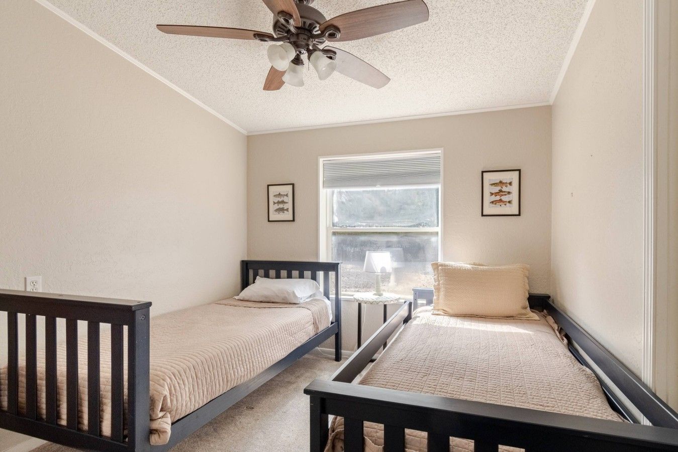 A bedroom with two single beds, a ceiling fan, a window with blinds, and framed pictures on the wall.