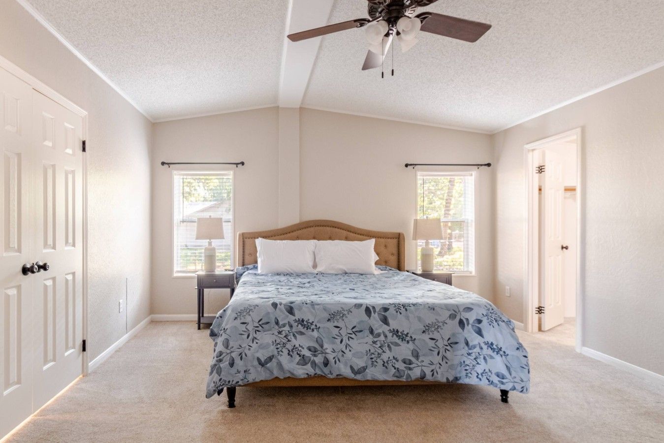 Une chambre à coucher comprenant un lit, deux tables de chevet, un ventilateur de plafond et deux fenêtres.