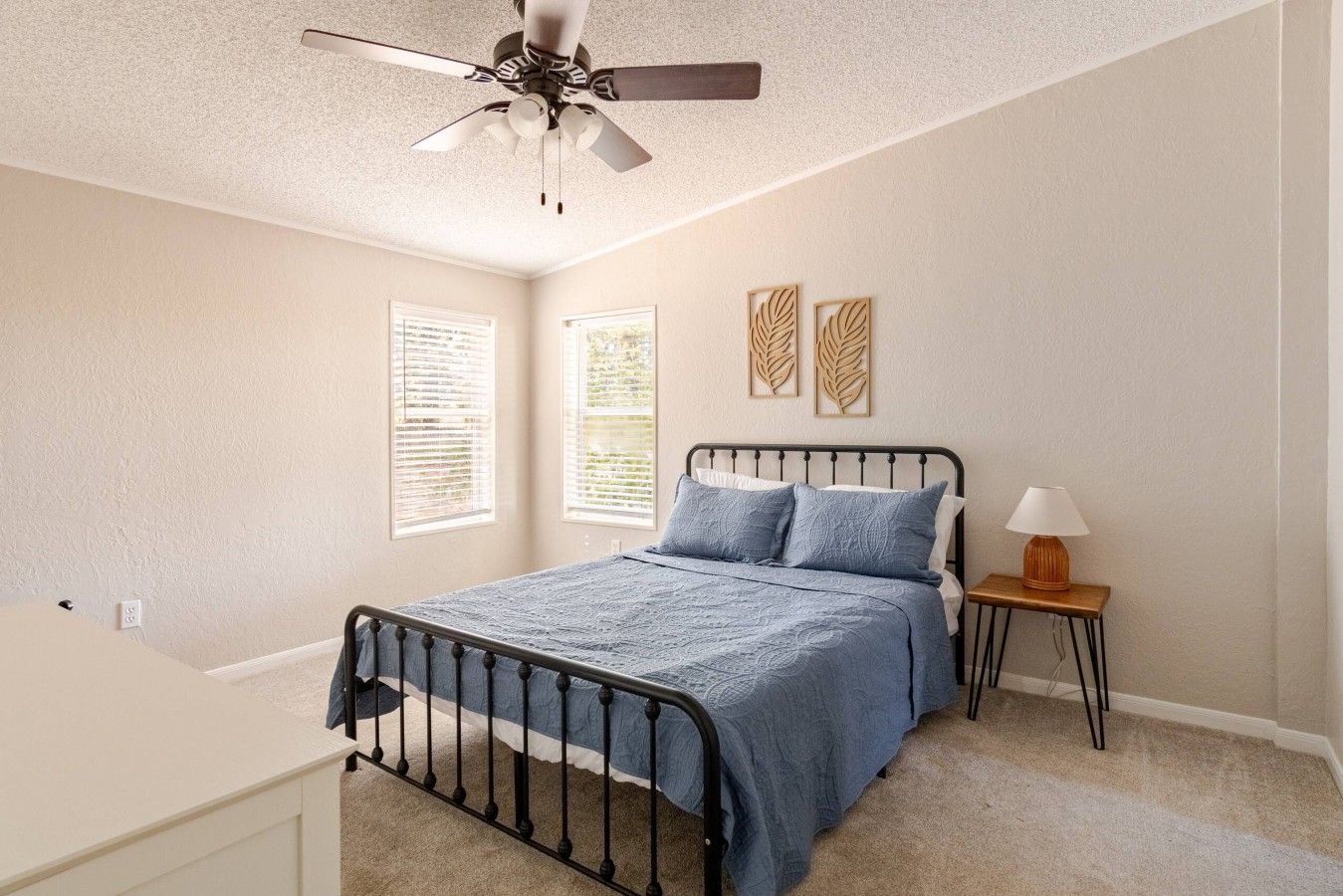 Une chambre à coucher comprenant un lit, une table de chevet, une lampe, un ventilateur de plafond et deux fenêtres équipées de stores.