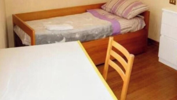 Een kleine kamer met een enkel bed, een houten stoel, een witte tafel en een wit dressoir.