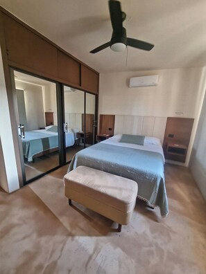 Ein Schlafzimmer mit Bett, einem Hocker, einem Deckenventilator, einem Spiegel und einer Klimaanlage.
