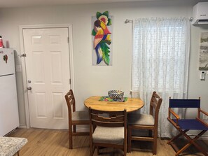 A dining area with a round wooden table, four wooden chairs, a colorful painting on the wall, a white door, and a window with patterned curtains. - Emma's Casa Bonita; Cozy Coastal Cottage Near Rockport Attractions (Rockport)