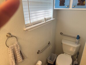 Ein Badezimmer mit Toilette, Handläufe, einem Handtuchhalter, einem Fenster mit Jalousien und einer kleinen Mülltonne.