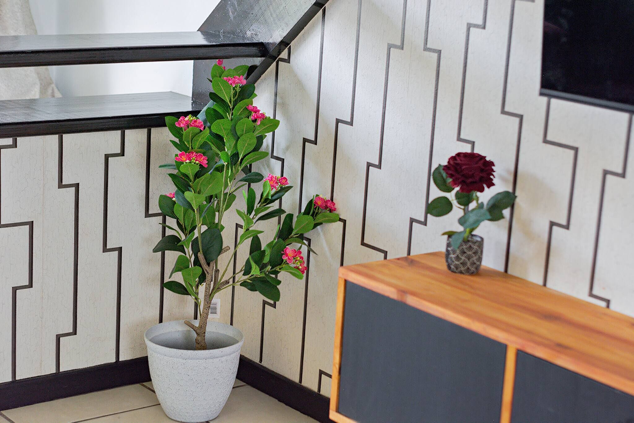 A potted plant with pink flowers, a single red rose, and a wooden side table in a corner with a zigzag patterned wall.