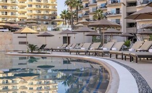Zona de piscina con sillones reclinables y sombrillas, rodeada de edificios altos.