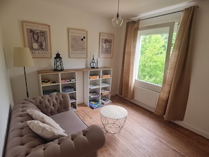 A living room with a sofa, a small round table, shelves with board games, and artwork on the wall. - Charming house with garden, 10 mm walk from the port and close to the beaches. (Saint-Quay-Perros)