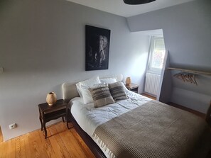 A bedroom with a bed, nightstands, a wall-mounted artwork, a window with blinds, and a radiator. - Charming house with garden, 10 mm walk from the port and close to the beaches. (Saint-Quay-Perros)
