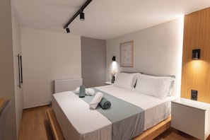 A modern bedroom with a bed, bedside tables, a radiator, and a wall-mounted light fixture. - Ground Living - Apartment Two (Volos)