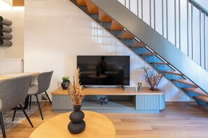 A modern living room with a TV, wooden flooring, grey chairs, a round wooden table, and a staircase. - Ground Living - Apartment Two (Volos)