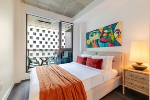 Una habitación de hotel con cama, un cuadro, una ventana que da a edificios y un balcón con sillas.