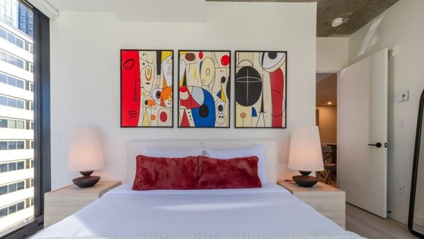 A hotel room with a large bed, two bedside lamps, abstract wall art, and a view of a building outside. - Modern Downtown Stay | Pool & Skyline (Montreal)