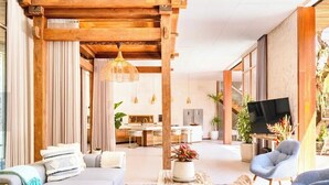 Ein modernes Wohnzimmer mit Holzbalkendecke, einer Couch, einem Sessel, einem Fernseher und einem Essbereich im Hintergrund.