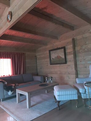 A wooden cabin interior with a sofa, a coffee table, a chair, and a painting on the wall.