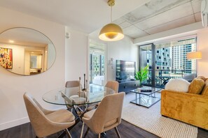 A modern living room with a glass dining table, beige chairs, a sofa, and a large window offering a city view. - Stylish Downtown Retreat | Pool & Views (Montreal)