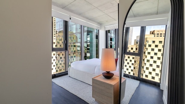 Ein modernes Hotelzimmer mit einem großen Bett, einer Nachttischlampe, einem Spiegel und Blick auf die Stadt durch große Fenster.