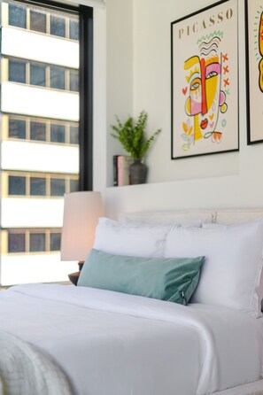 A bed with white bedding and a teal pillow, a wall-mounted lamp, and a framed Picasso artwork. - Premium Downtown Living | Pool Gym & Views (Montreal)