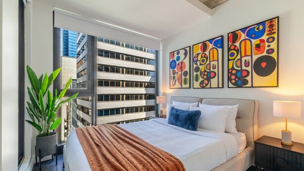 Quarto de hotel com cama, janela ampla, planta em vaso e arte abstrata na parede.