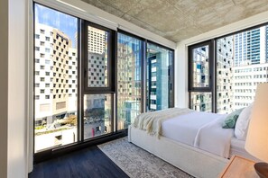 Quarto de hotel moderno com janelas amplas, uma cama e vista para a paisagem urbana.