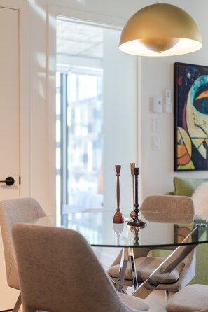 Ein moderner Essbereich mit einem Glastisch, zwei Stühlen, einer Pendelleuchte und einem Gemälde an der Wand.