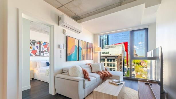 Ein modernes Wohnzimmer mit Sofa, Couchtisch und Fernseher, großen Fenstern mit Blick auf die Stadt und Kunstwerken an den Wänden.