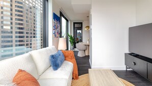 Ett modernt vardagsrum med en soffa, en tv och utsikt över en stadssiluett genom stora fönster.