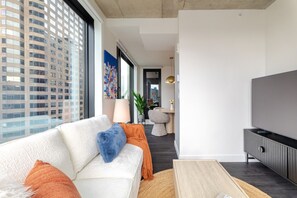 A modern living room with a sofa, TV, and a view of a cityscape through large windows. - Luxe Central Retreat | Free Parking & Pool (Montréal)