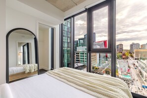A bedroom with a large bed, a mirror, and a city view through large windows. - Luxe Central Retreat | Free Parking & Pool (Montréal)